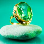 A green sapphire ring with an intricate gold setting was placed on a smooth grey stone against a vibrant green background.