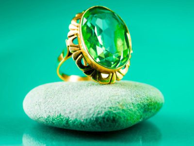 A green sapphire ring with an intricate gold setting was placed on a smooth grey stone against a vibrant green background.