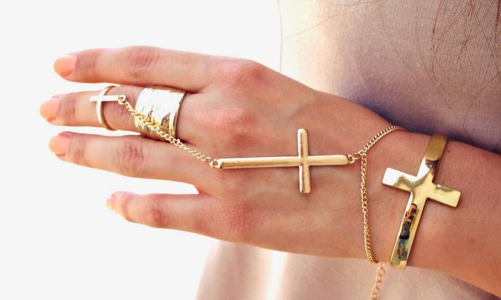 A hand with a cross ring connected by a chain to a cross bracelet.