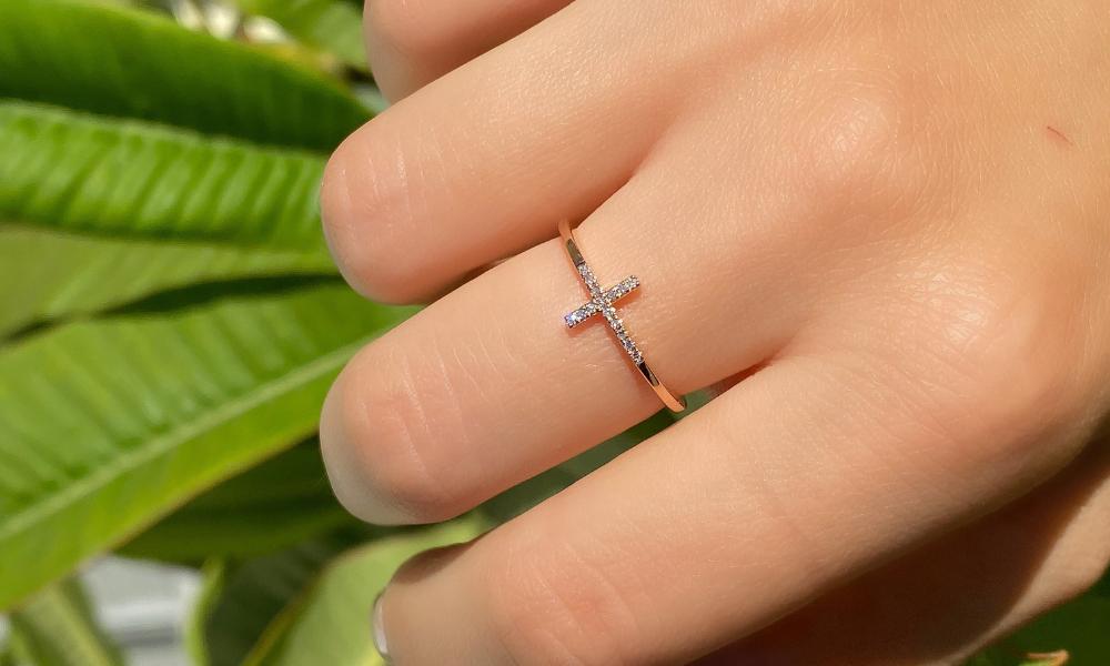 A hand wearing a diamond cross ring over a background of green leaves.