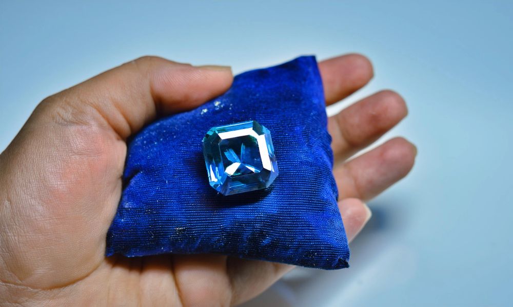 A hand holds a blue fabric with a large, faceted blue emerald on it.