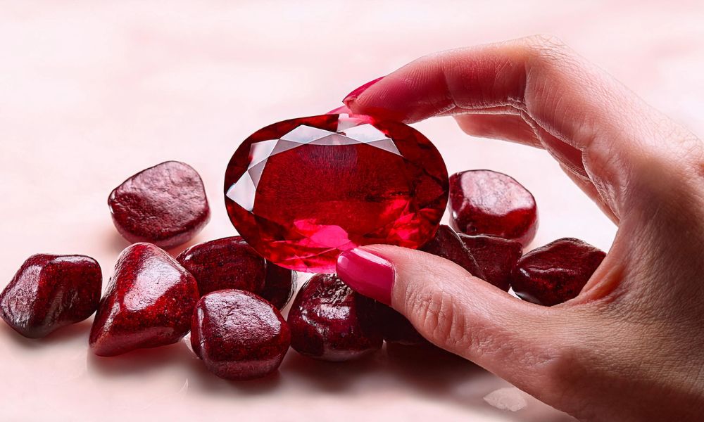 A hand with pink nail polish holds a large, faceted ruby above smaller garnets.