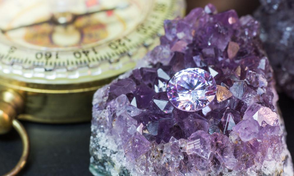 A round-cut purple diamond atop an amethyst cluster with a blurred compass in the background.