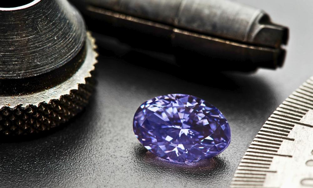 A sparkling purple diamond next to a metal gauge and jeweler's tools on a dark surface.