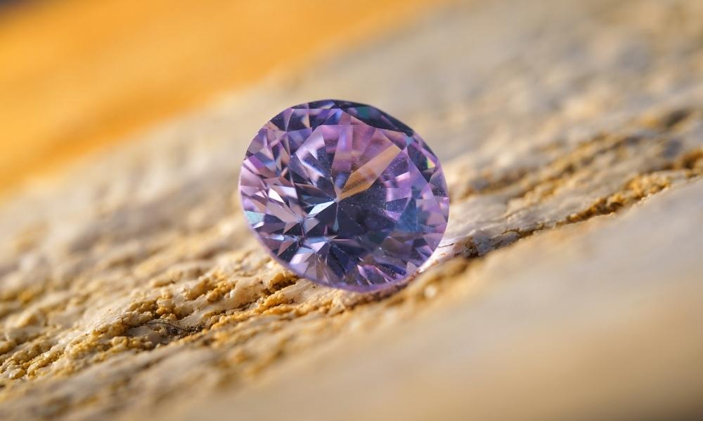 A sparkling purple diamond placed on a textured, sandy surface.