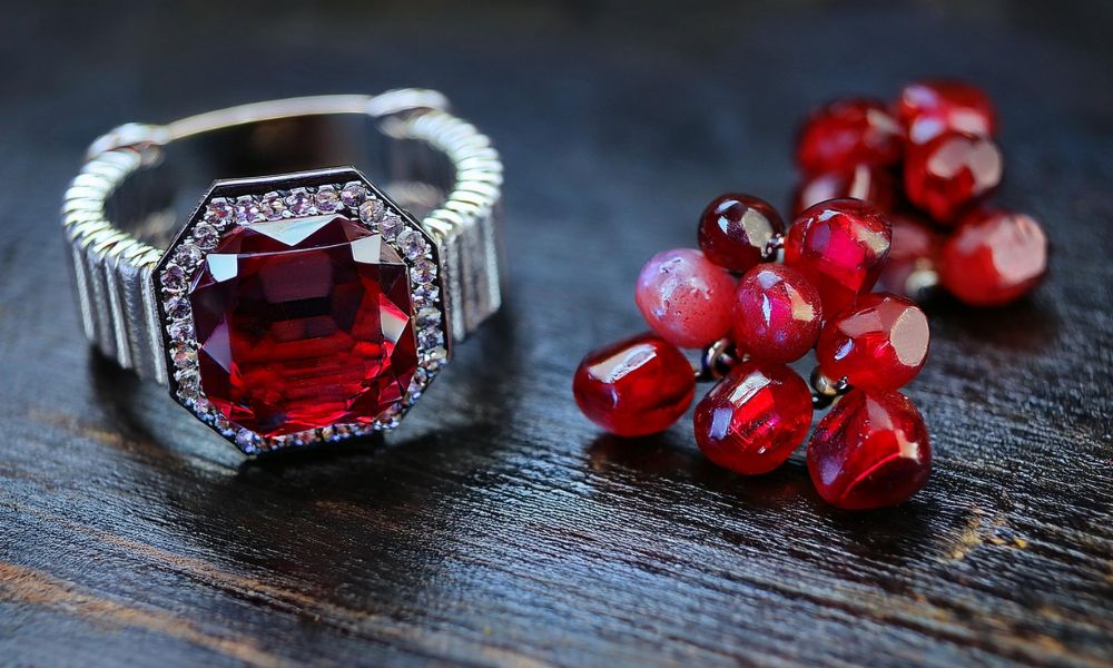 A large red gemstone ring next to a cluster of shiny red beads on a wooden surface.