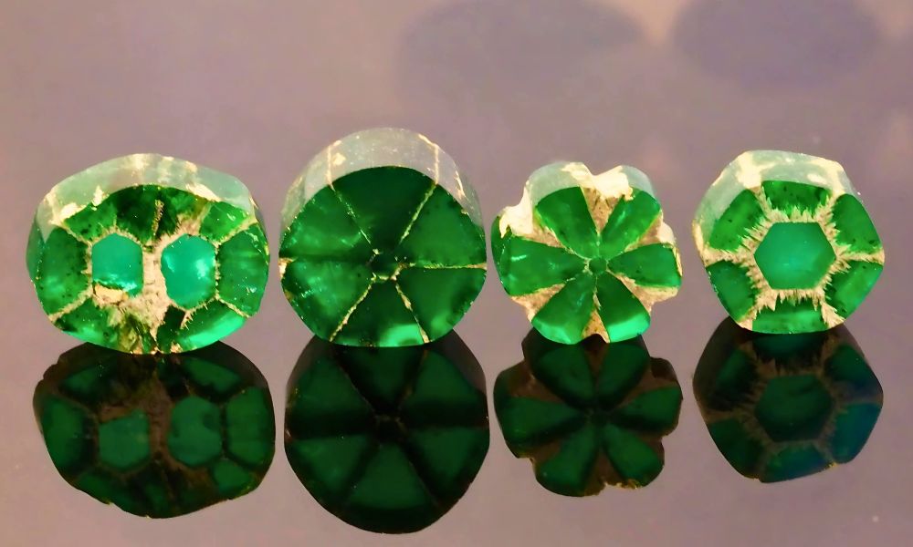 Four trapiche emerald with intricate cut patterns, displayed on a reflective surface.