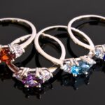 Four gemstone rings with red, purple, and blue stones on a reflective black surface.