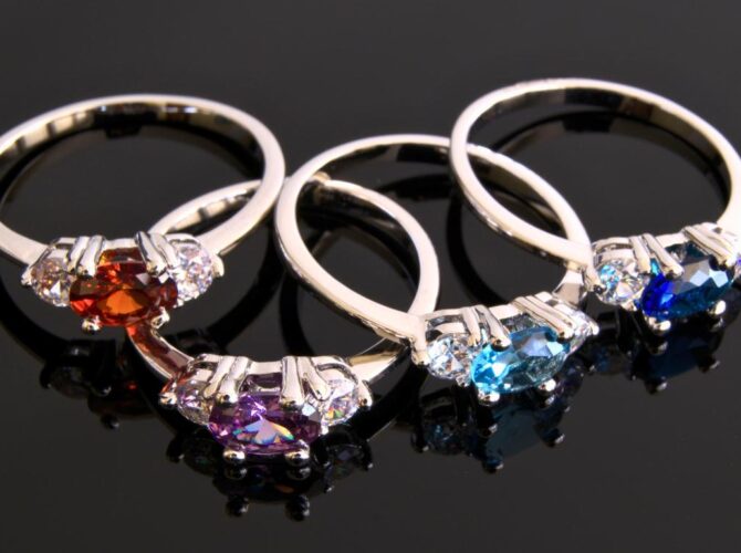 Four gemstone rings with red, purple, and blue stones on a reflective black surface.