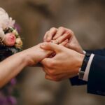 A close-up of hands exchanging wedding gemstone rings during a wedding ceremony, with a bouquet in the background.