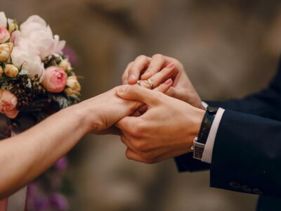 A close-up of hands exchanging wedding gemstone rings during a wedding ceremony, with a bouquet in the background.