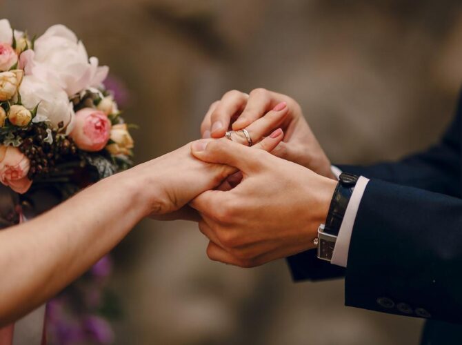 A close-up of hands exchanging wedding gemstone rings during a wedding ceremony, with a bouquet in the background.