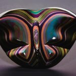 A rainbow obsidian, symmetrical glass sculpture with a reflective, wavy design on a gray background.