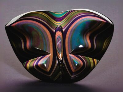 A rainbow obsidian, symmetrical glass sculpture with a reflective, wavy design on a gray background.