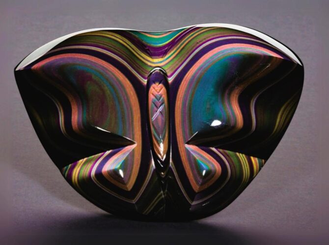 A rainbow obsidian, symmetrical glass sculpture with a reflective, wavy design on a gray background.