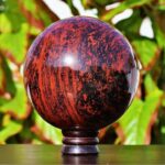 A polished red obsidian on a stand with green foliage in the background.