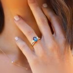 A close-up of a hand with a stunning blue gemstone engagement rings and diamonds, resting on a woman's neck. Soft, natural lighting.