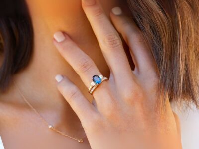 A close-up of a hand with a stunning blue gemstone engagement rings and diamonds, resting on a woman's neck. Soft, natural lighting.