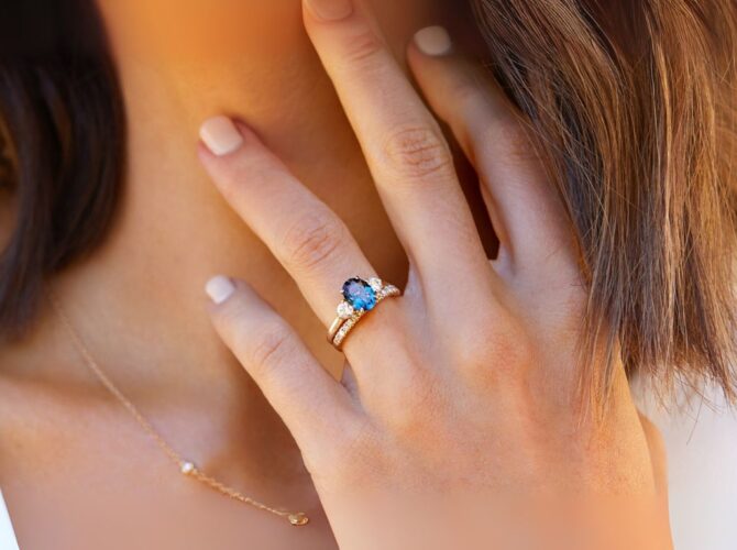 A close-up of a hand with a stunning blue gemstone engagement rings and diamonds, resting on a woman's neck. Soft, natural lighting.