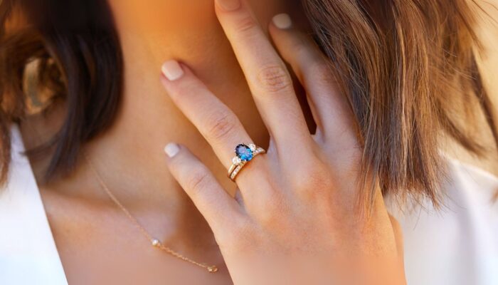 A close-up of a hand with a stunning blue gemstone engagement rings and diamonds, resting on a woman's neck. Soft, natural lighting.